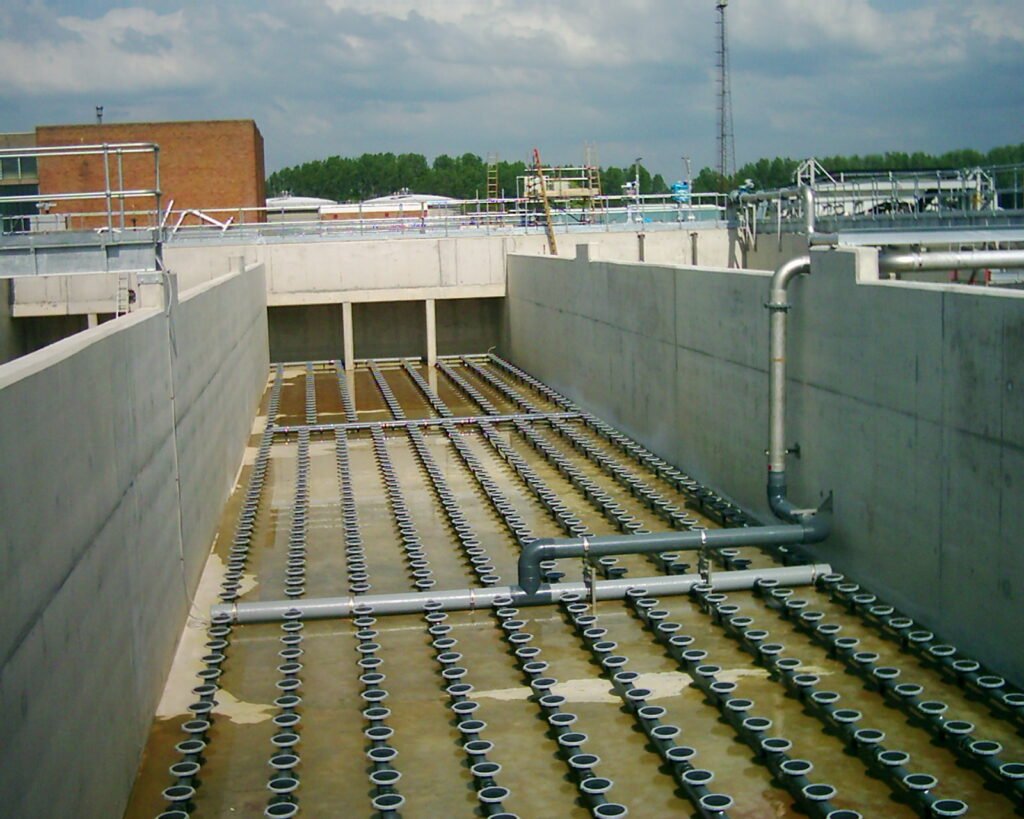 Concrete tank with aeration diffusers in a wastewater treatment plant, showing pipes and infrastructure under a cloudy sky.