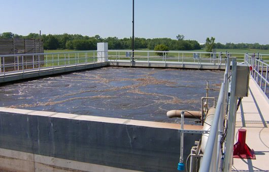 Wastewater treatment tank with aeration bubbles, surrounded by fencing, located outdoors with trees in the background.