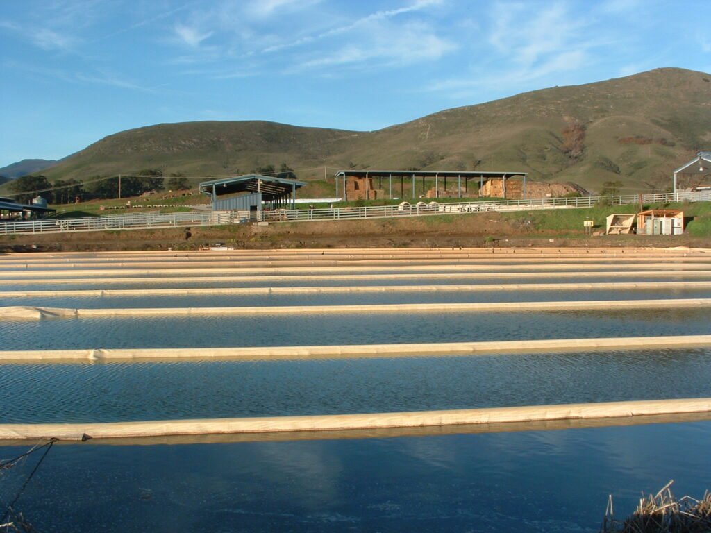 Aquaculture facility with water-filled linear tanks, situated in a rural area with green hills and blue sky in the background.