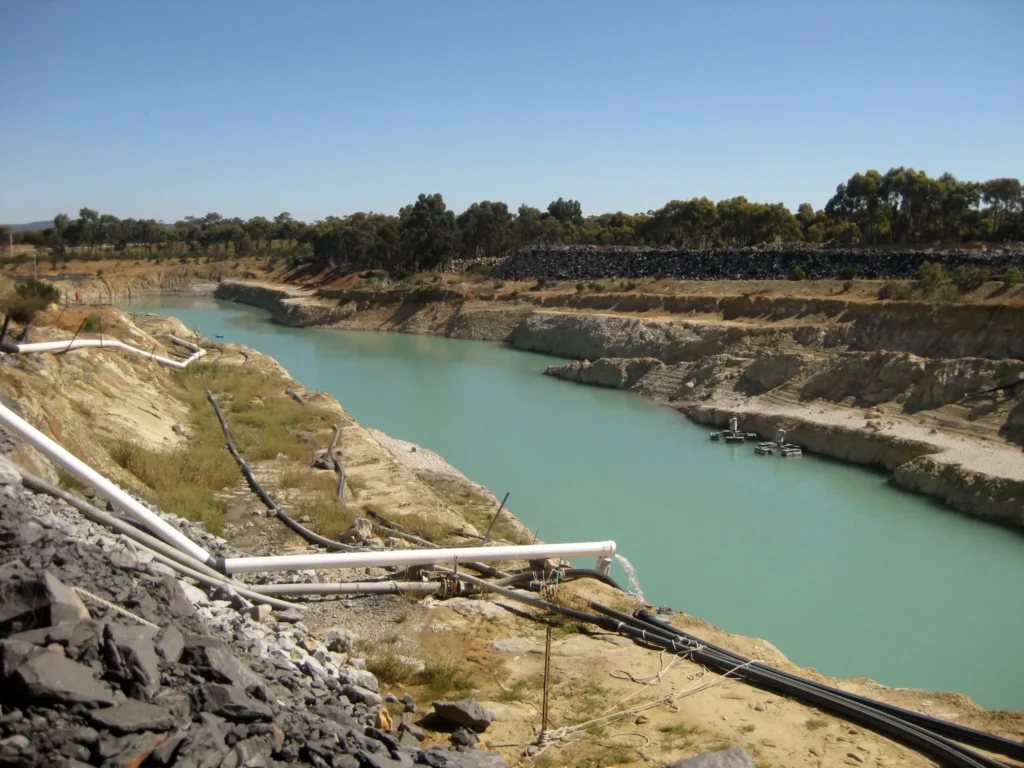 Aquamarine water in a mining pit with surrounding rocky terrain and water pipes along the edges, under a clear blue sky.