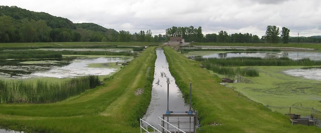 View of a wastewater treatment facility featuring grassy pathways, water channels, and surrounding trees under a cloudy sky.