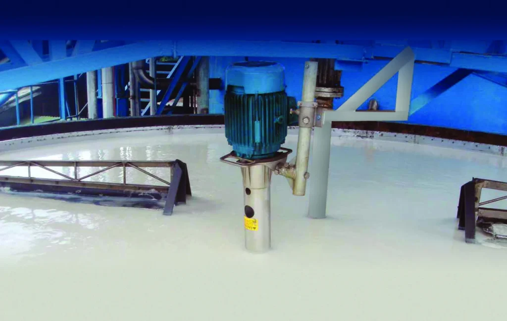 Industrial mixing tank with a blue motor and metal structures, mixing a white liquid; background shows blue structural supports.