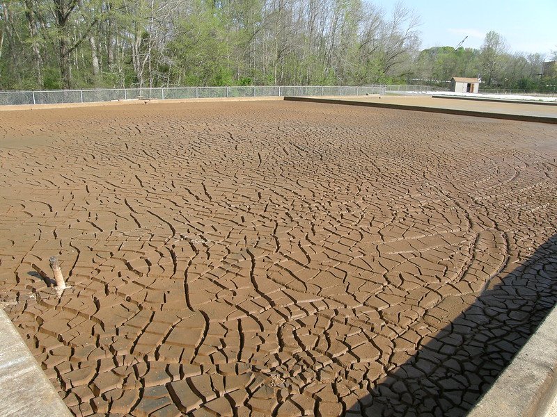 A concrete basin filled with cracked, dried mud, surrounded by a chain-link fence and trees in the background.