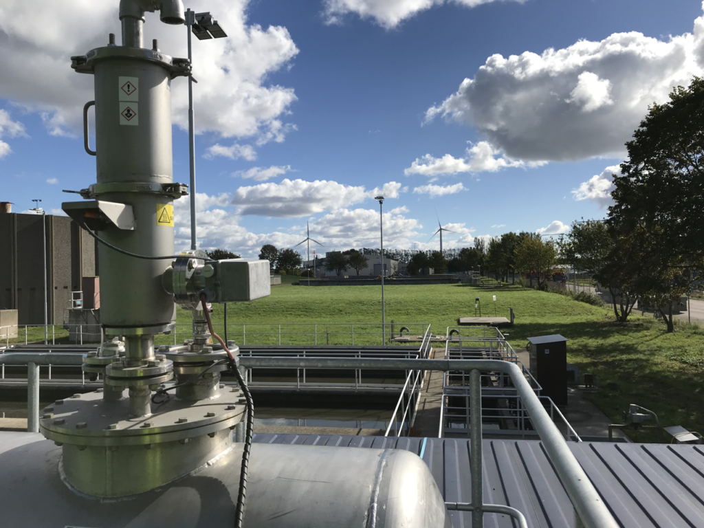 Industrial facility with metal equipment in the foreground, grassy field, and wind turbines under a cloudy blue sky.