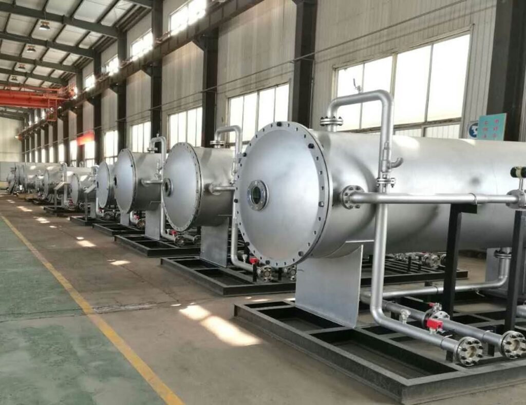 Row of large industrial pressure vessels in a factory setting with metal pipes and fittings, under a high ceiling.