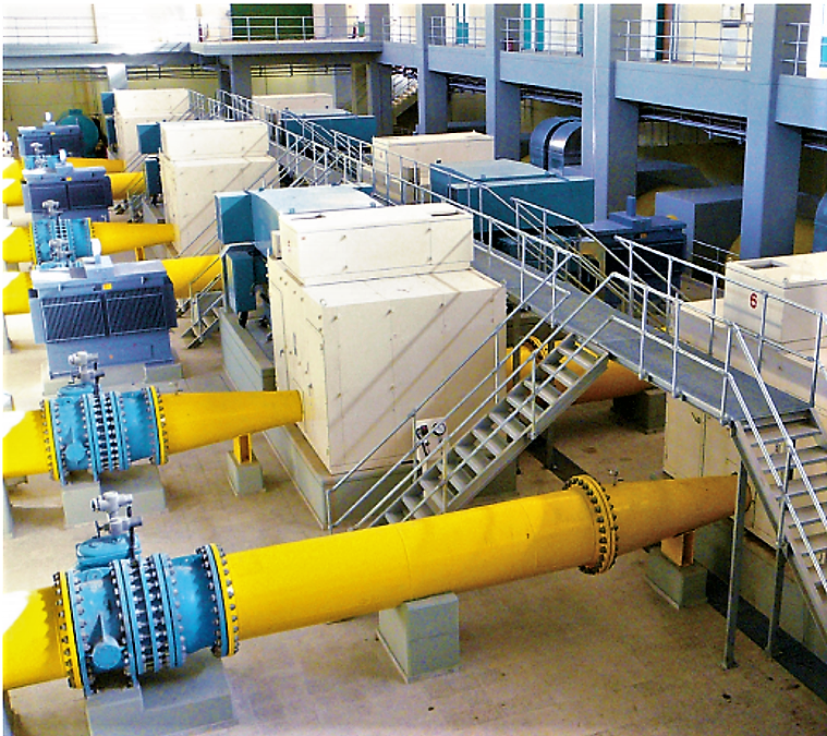 Industrial facility interior with large yellow pipes, blue valves, and machinery connected by metal walkways and stairs.