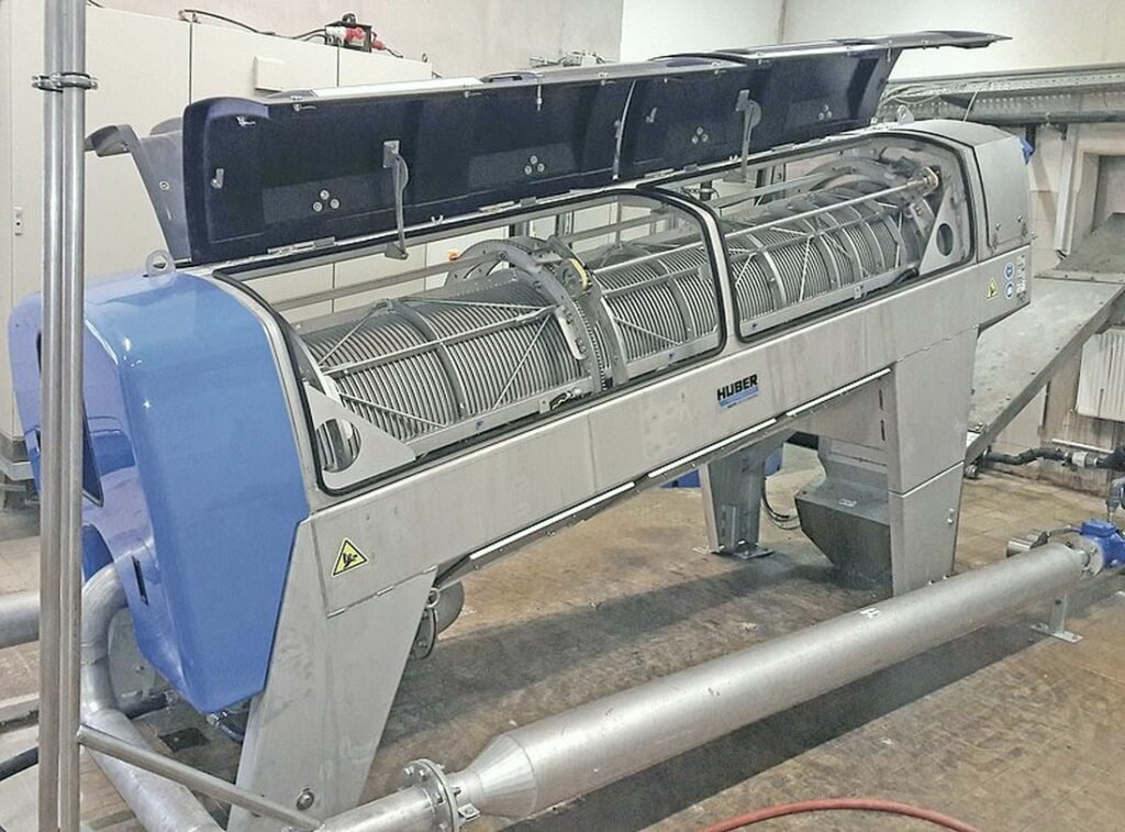 Industrial stainless steel screw press machine in a factory setting, with open top and visible internal components.