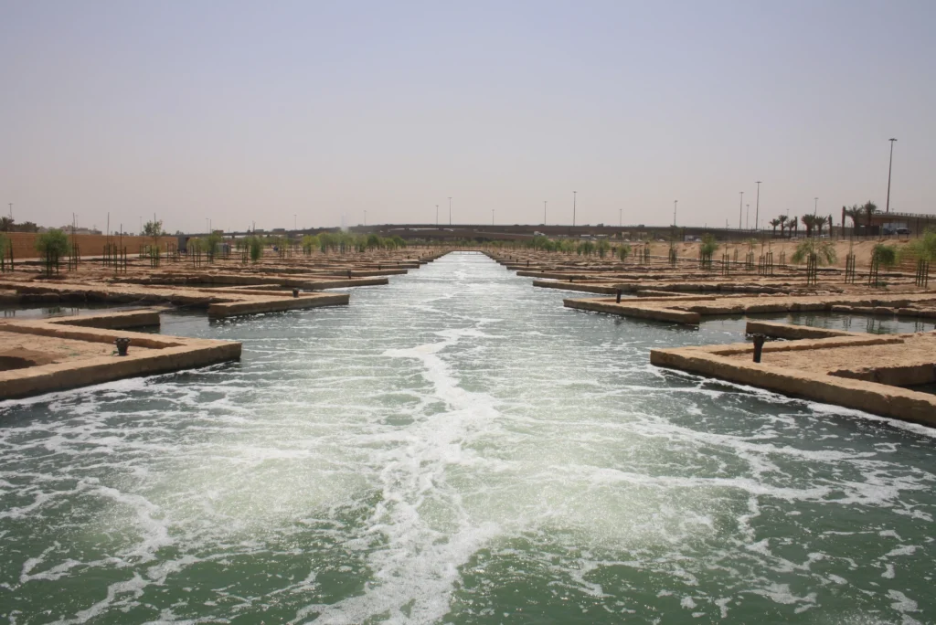 A large rectangular fish farming pond features small bubble aerators creating ripples and bubbles on the water's surface. The pond is lined with concrete walkways, and small trees are visible along each side under a clear blue sky.