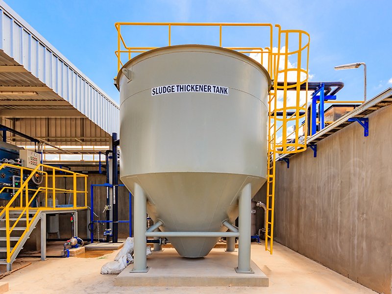 Large cylindrical sludge thickener tank at an industrial facility, surrounded by yellow-railed walkways and blue piping.