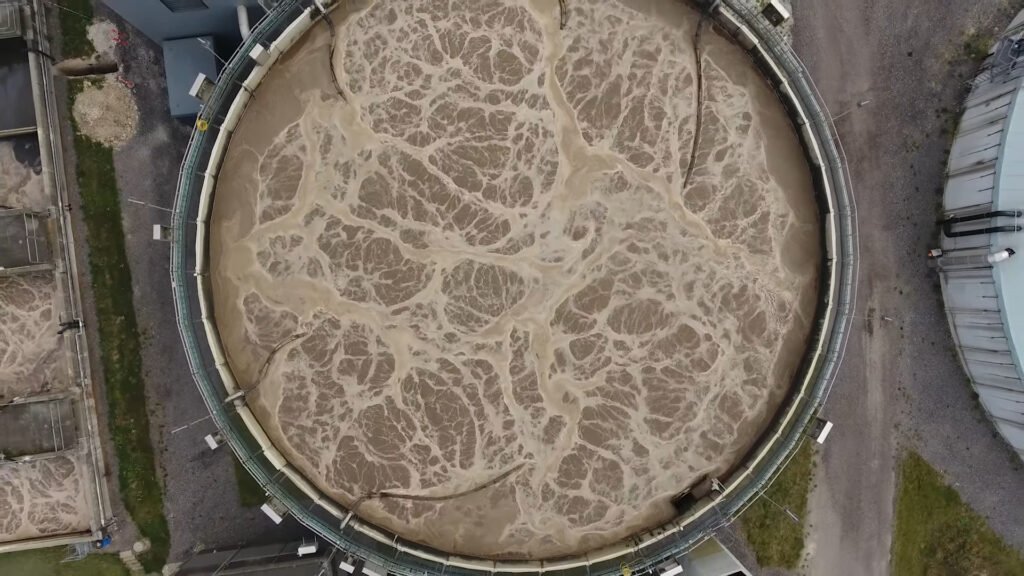 Aerial view of a large circular tank at a wastewater treatment plant with swirling water patterns inside.