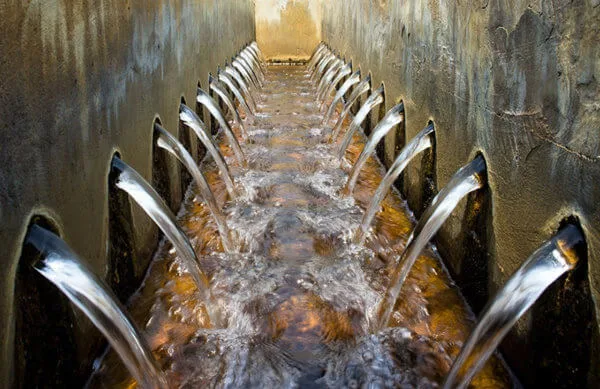 Water flows from multiple spouts in parallel rows along the walls of a rectangular channel, creating a symmetrical pattern.