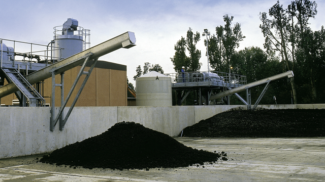Industrial facility with large piles of compost, conveyor belts, storage tanks, and trees in the background.