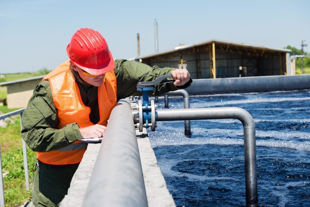 A worker in a red helmet and orange safety vest adjusts a valve at an outdoor water treatment facility.