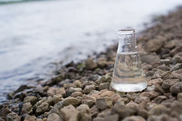 A glass Erlenmeyer flask filled with clear water sits on rocky ground near the edge of a natural body of water, evoking ideas of biomimetic water purification with the shoreline and reflective surface visible in the background.