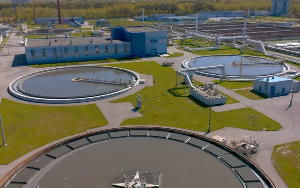 Aerial view of a wastewater treatment plant with several large circular settling tanks, part of H2O Innovation’s advanced water treatment operations, connected by pipes and surrounded by green grass and industrial buildings in the background.
