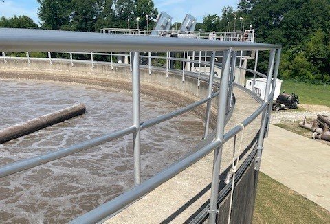 A large circular waste water treatment tank with metal railings, filled with bubbling water, is set outdoors at the Arlington Treatment Plant near grass, trees, and a paved path.