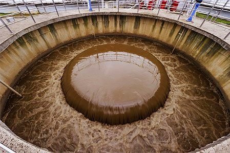 An aerial view captures a circular wastewater treatment tank, where brown water swirls around a central opening. This outdoor facility, surrounded by metal railings, showcases the process of transforming wastewater into valuable resources.