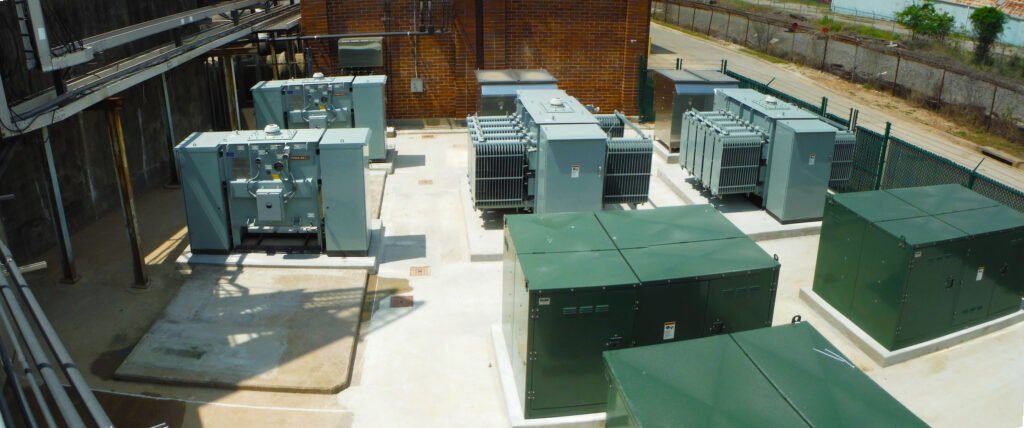 View of an electrical substation on 69th Street with multiple green and gray transformers and equipment on a concrete platform. Fenced area, with a brick building and road in the background. Sunny weather casts shadows on the ground.