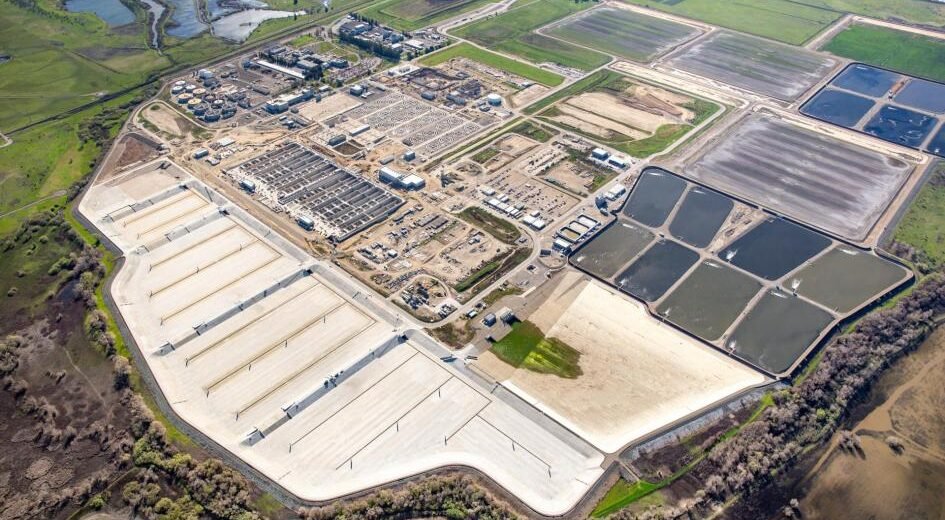 An aerial view of the Sacramento water treatment facility reveals multiple rectangular ponds and large building structures. This regional site is surrounded by green fields and wetlands, with residential areas visible in the distant background, highlighting its role in effective wastewater treatment.