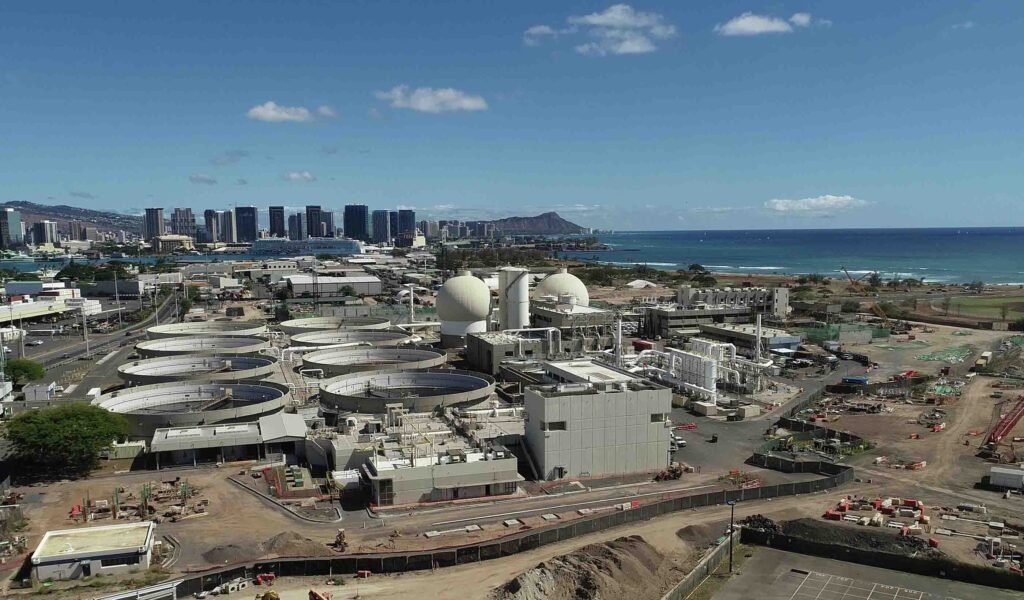 Aerial view of a large wastewater treatment plant near a coastline, showcasing multiple circular tanks and infrastructure. Sand Island's city skyline and mountain range grace the background under a clear blue sky.