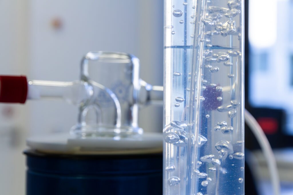Close-up of a laboratory setup featuring a glass condenser connected to a flask. Clear liquid with bubbles is flowing through a vertical tube on the right, suggesting an advanced oxidation process, against a blurred background.