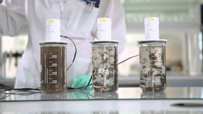A person in a lab coat holds a clipboard, observing three beakers filled with brown liquid and sediment, each equipped with a stirring device. The presence of magnetic nanoparticles implies advanced water treatment in the blurred laboratory setting.