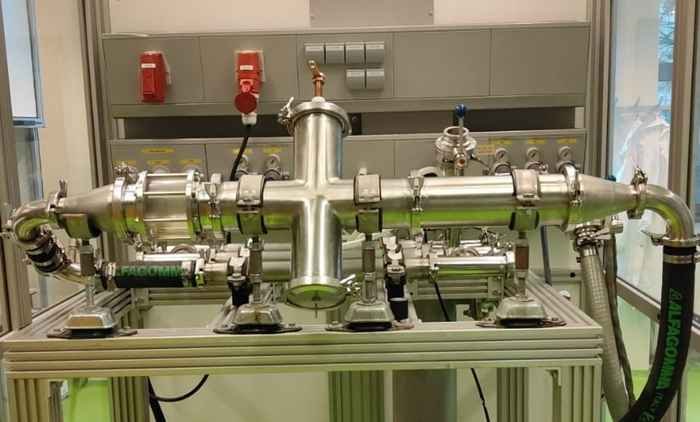 A complex metal piping system installed on a platform, featuring intersecting pipes and various valves, connectors, and catalytic gauges. The background includes a panel with emergency buttons and additional fittings on a control board. This setup is integral to the plasma-assisted process.