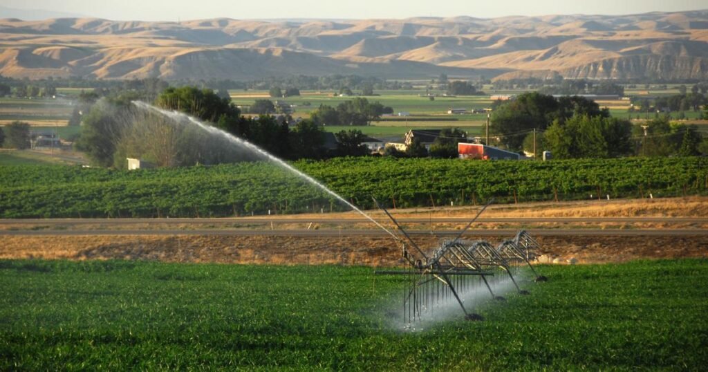 Irrigation system watering green fields with a backdrop of farms, trees, and rolling hills in the distance.