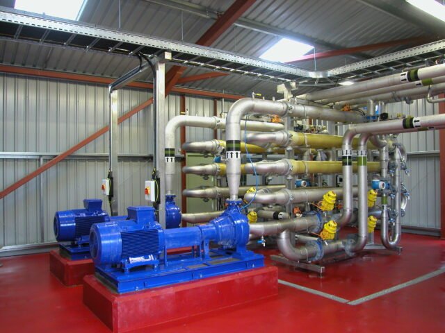 Industrial piping system with blue water pumps, stainless steel pipes, and yellow valves inside a metal-walled facility.