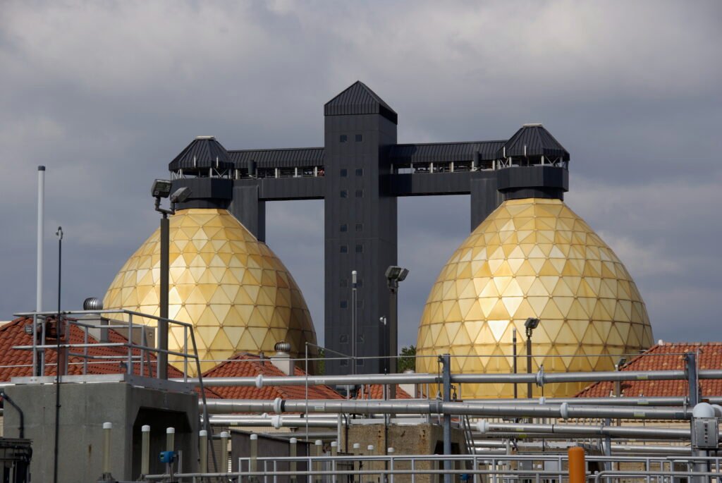 Two large, golden geodesic dome structures stand beside a dark, rectangular tower at the Back River Wastewater Treatment Plant, surrounded by industrial piping and buildings under a cloudy sky.