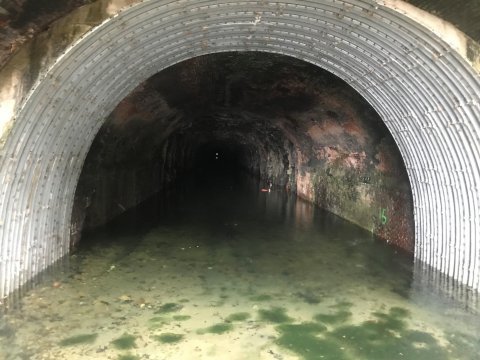 A narrow, water-filled tunnel with a rounded, corrugated metal entrance. The tunnel walls are damp and mossy, indicative of water pollution control efforts. Inside, the murky water is filled with scattered debris as light fades into darkness.