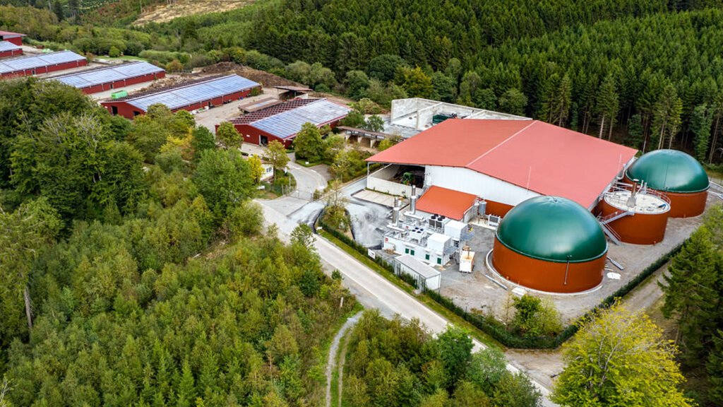 Aerial view of a biogas plant with large storage tanks and solar panel-covered buildings surrounded by lush green forest.