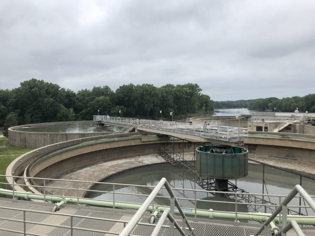 View of a wastewater treatment facility with circular tanks, metal walkways, and railings, set against a backdrop of green trees and a cloudy sky in Des Moines.