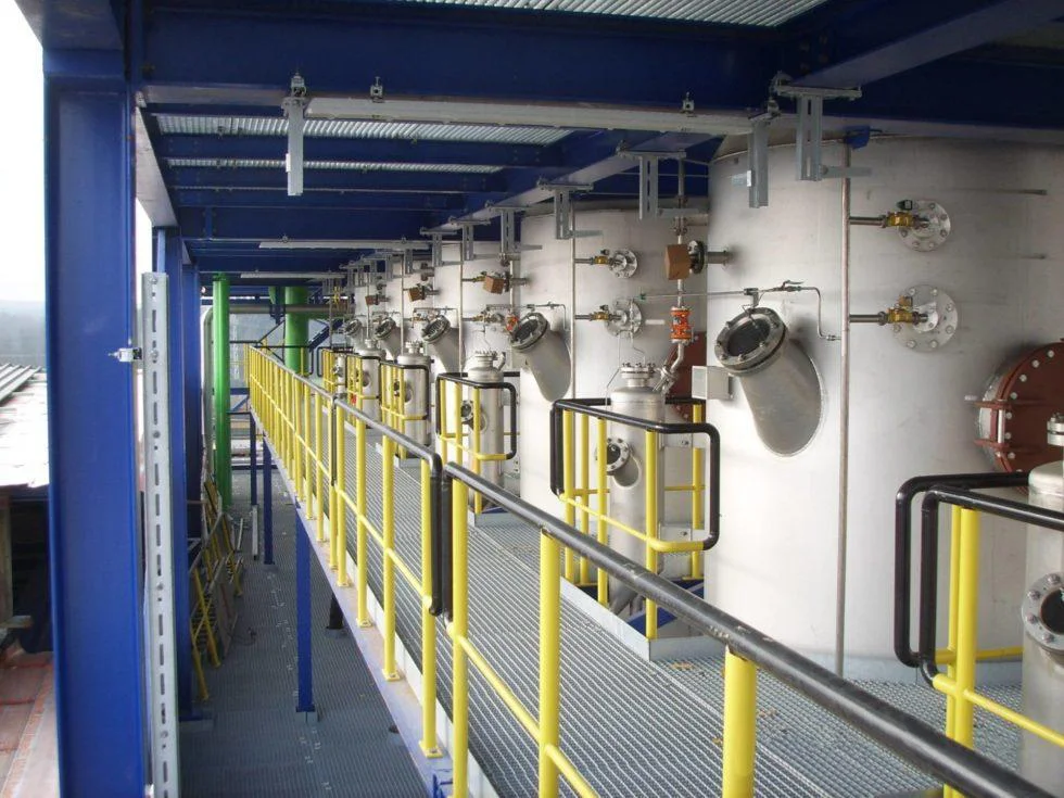 The industrial facility interior features a row of large, connected cylindrical tanks along a wall. A yellow-railed metal walkway runs parallel to these crystallization tanks. Pipes and valves are visible, with overhead metal beams supporting the structure, emphasizing its role in fluidized bed water treatment.