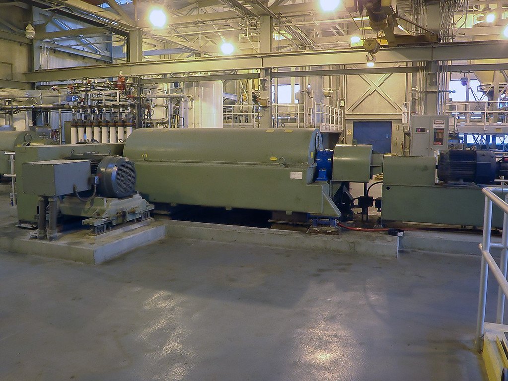 Industrial machinery inside a large facility in Essex County showcases a long, green metal cylinder connected to various components. Overhead lights illuminate the space, highlighting pipes and equipment, reminiscent of those used to manage sewer overflow systems.