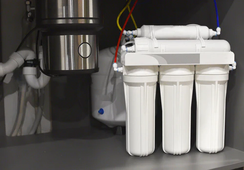 Under-sink water purification system featuring three white cylindrical filters and a metallic disposal unit. Color-coded pipes for water flow connection are visible. This reverse osmosis setup is installed in a gray cabinet, ensuring efficient filtering for your home.