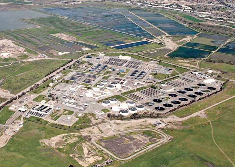 Aerial view of San Jose Creek wastewater treatment plant, showcasing numerous circular and rectangular tanks amidst lush greenery. Nearby, several rectangular ponds enhance water reclamation efforts. Roads and some buildings can be seen in the upper right corner.