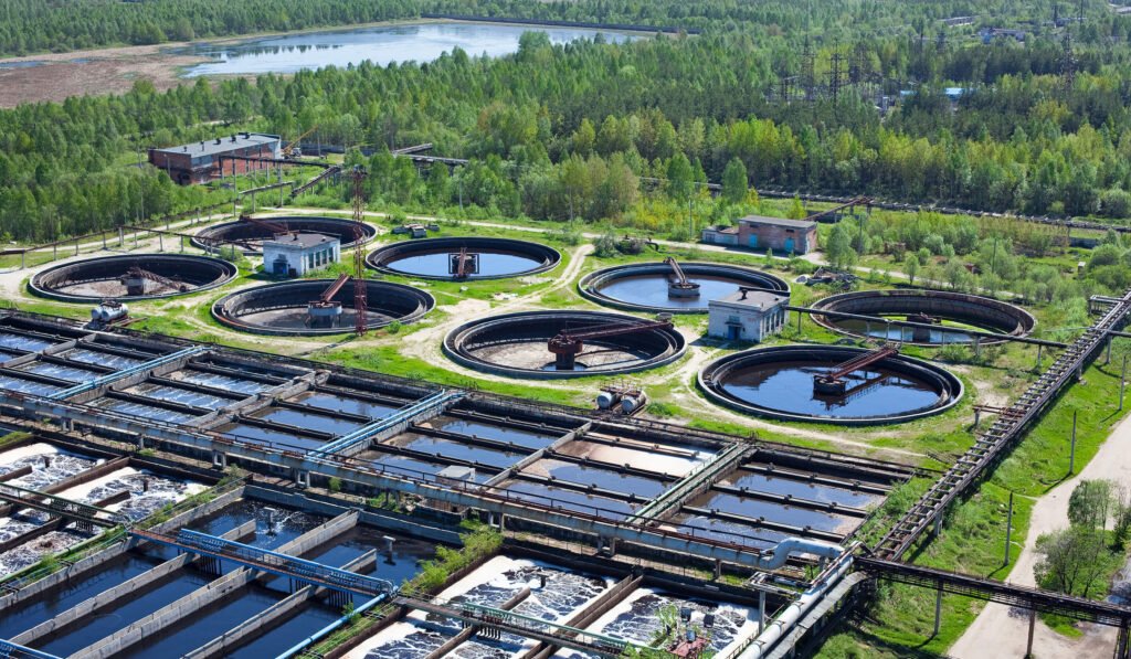 Aerial view of a wastewater treatment plant with multiple circular settling tanks and rectangular filtration pools surrounded by trees.