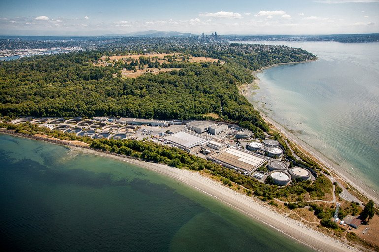 Aerial view of the West Point Wastewater Treatment facility near a coastline. The plant is surrounded by trees and bordered by a sandy beach and calm waters, with a city skyline visible in the distance under a clear blue sky.