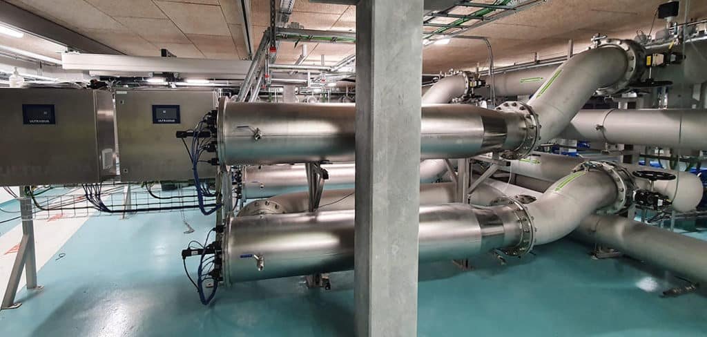 Industrial facility with large stainless steel pipes and electronic control panels on a clean, blue-painted floor.