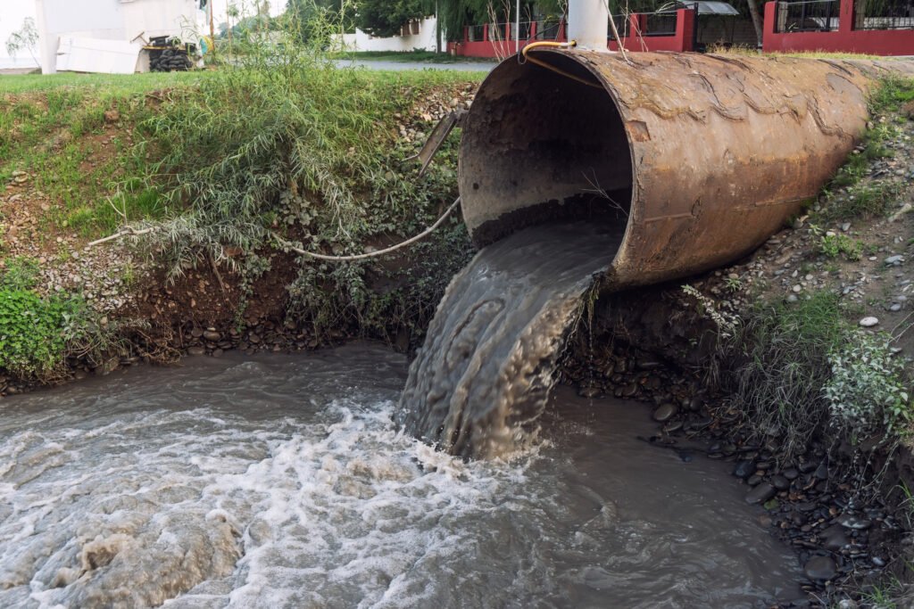 A large rusted pipe discharges polluted wastewater into a river, surrounded by vegetation and a rocky bank.