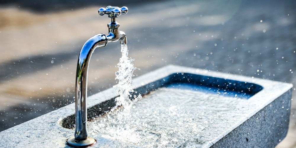 Water flows from a chrome faucet into a concrete basin outdoors, with droplets splashing in the sunlight.