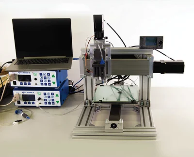 A 3D printer setup with a laptop and control units on a desk showcases the future of manufacturing. Equipped with smart materials, the printer boasts a metal frame and glass printing bed. Wires connect to control boxes, while a small screen is mounted on the frame for seamless operation.
