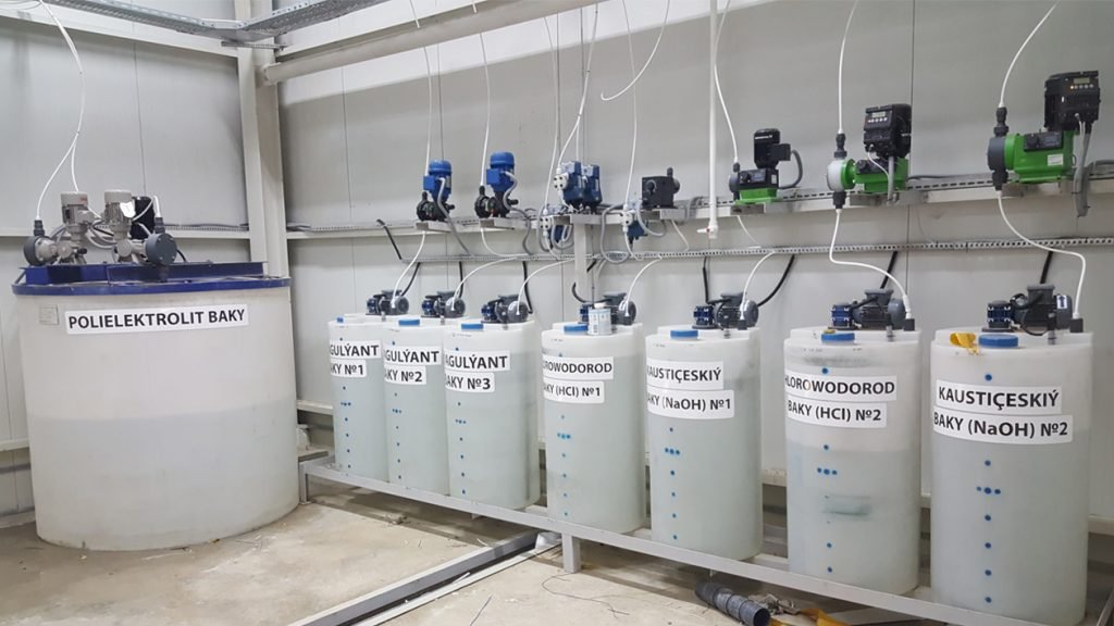A row of transparent tanks labeled with chemical names like HCl and NaOH, equipped with various pipes and valves, defines the clean industrial setting. A larger blue-lidded tank titled "Polielektrolit Baky" stands to the left, adjacent to a crucial chlorine pump for water in the system.