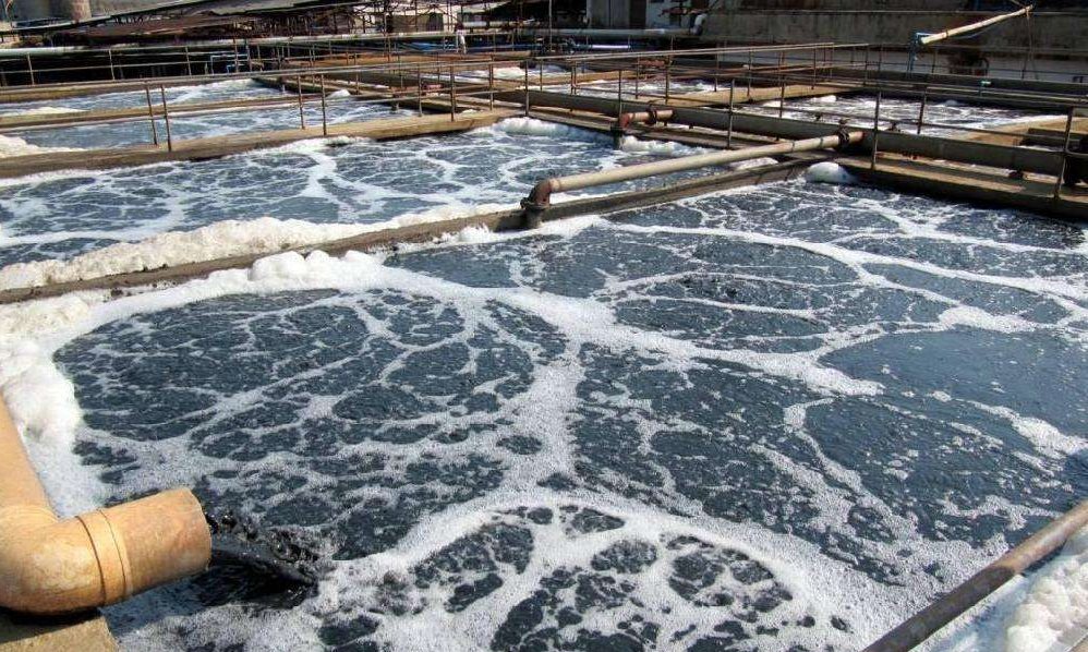 An industrial wastewater treatment facility features large open tanks filled with foamy, churning water. Pipes run along the edges, while a defoamer pipe in the foreground releases water into a tank. The environment appears industrial and functional.