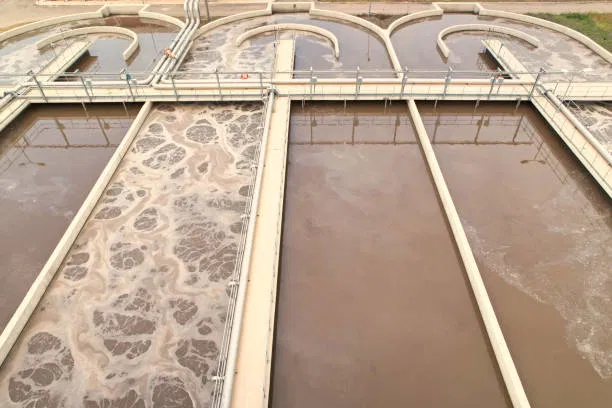 Aerial view of a wastewater treatment facility reveals various rectangular tanks, some brimming with aerated brown water treated with ferric chloride, while others remain clearer. The infrastructure features sturdy metal railings and gracefully curved water channels.