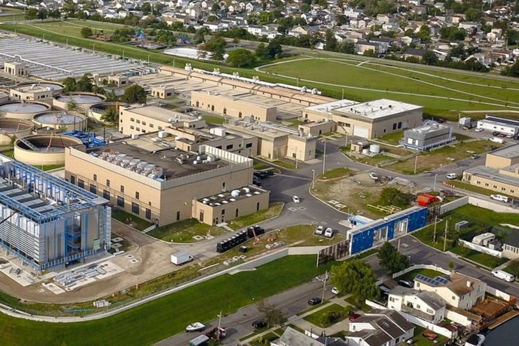 Aerial view of the Bay Park Sewage Treatment Plant, featuring several large buildings and circular structures. The facility is surrounded by greenery and residential neighborhoods in Nassau County. Roads and parking areas are clearly visible, integrating the plant within its vibrant community setting.