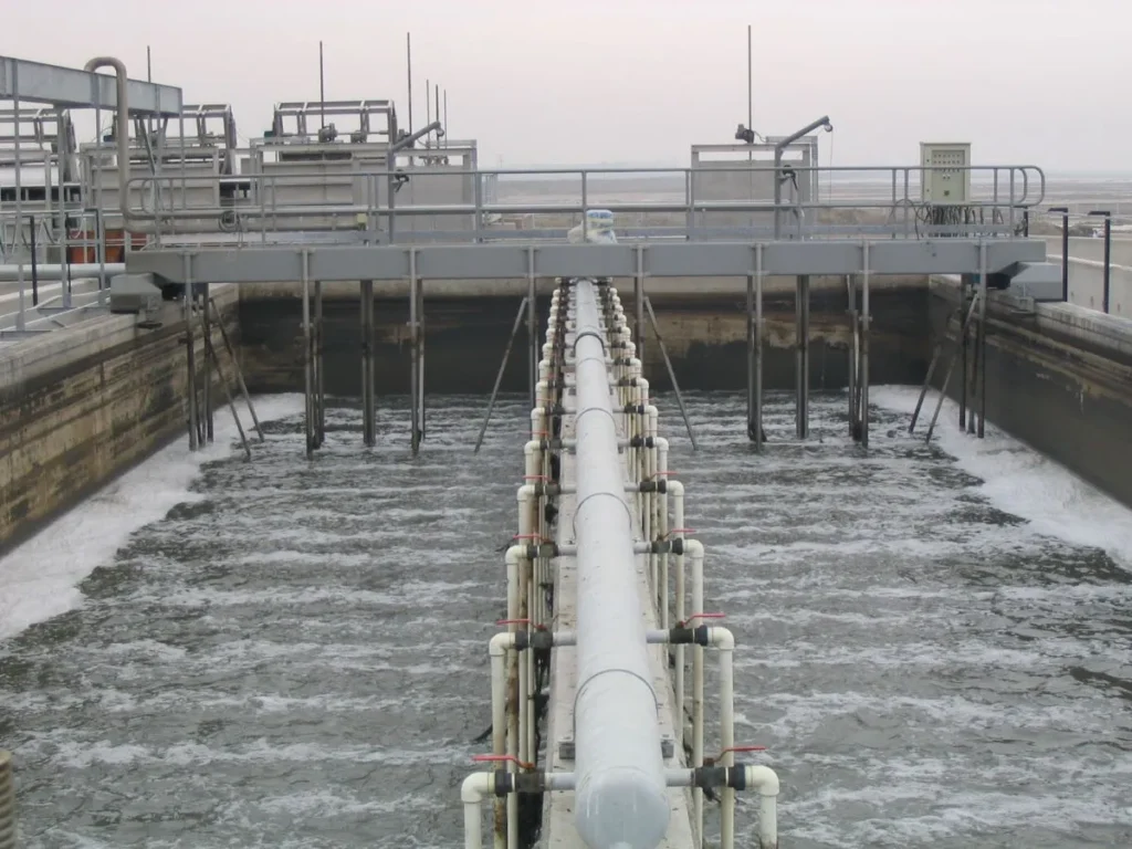 A large industrial wastewater treatment facility features a series of pipes and channels. Water flows below through the system's grit chamber, surrounded by metal structures and walkways. The overcast sky adds to the muted, industrial appearance.