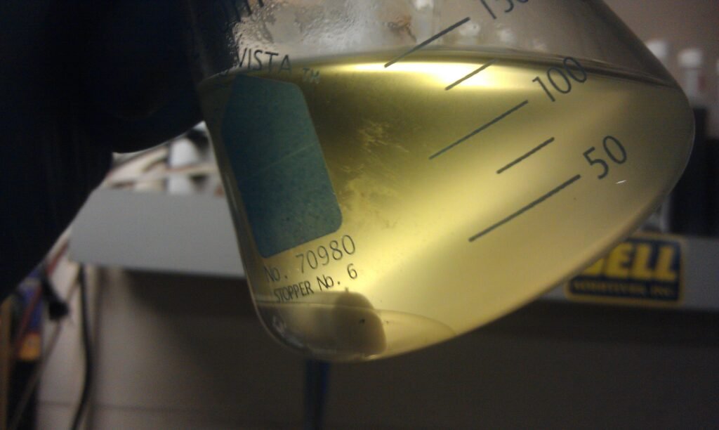 Hand holding a beaker filled with yellow liquid, marked with measurements up to 150 ml, in a laboratory setting.