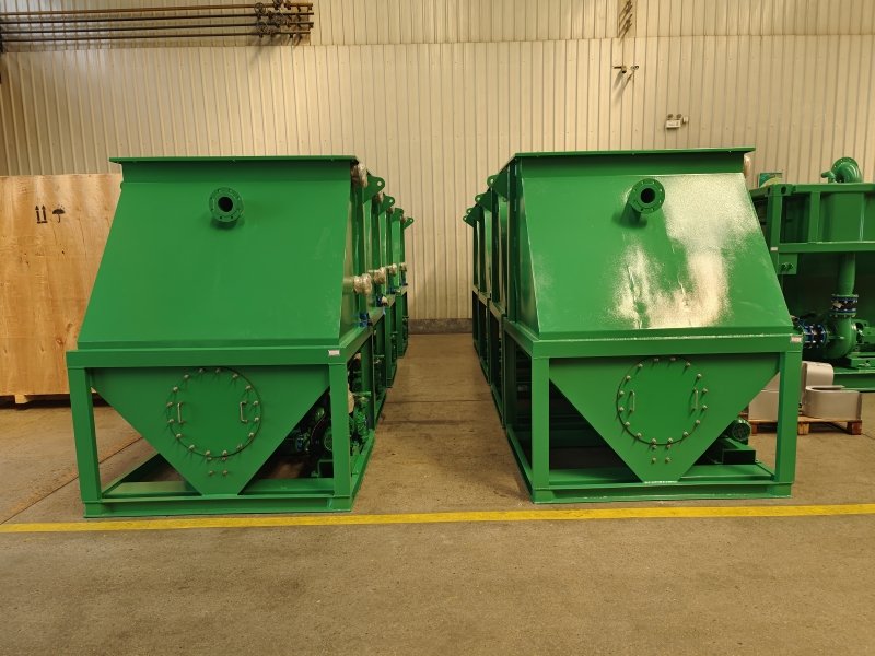 Two large green industrial machines, resembling inclined plate clarifiers, stand side by side in a warehouse. They feature circular access panels on the front and rest on a concrete floor, surrounded by a wooden crate and other equipment, likely used for efficient oil/water separation.